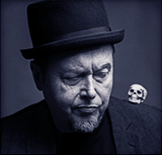 The man in the image have a short beard and a hat. On his shoulder rests a small skull. He looks concentrated. This is Peter Rosengren, the magician.